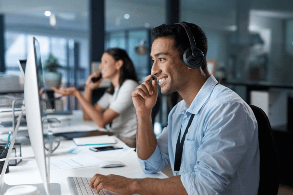 image of a man and a woman in the background talking on the phone using a headset. They're both looking at their flat screen computers and appear to be smiling.