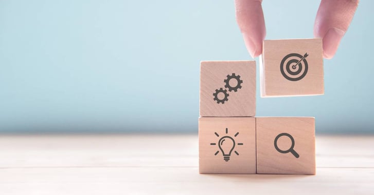 4 wooden blocks with icons of a target, cogs, light bulb, and magnifying glass, being stacked