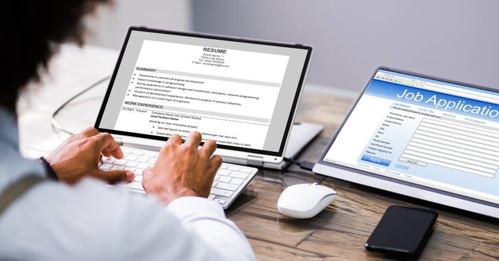 Man typing a resume on a laptop while looking at a job application 